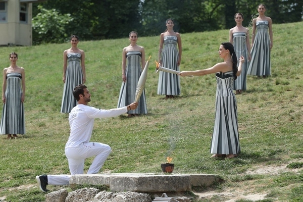 扮演最高女祭司的演員瑪麗·米娜（前右）點燃第一棒火炬手——希臘運動員、東京奧運會男子賽艇項目金牌得主斯特凡諾斯·恩圖斯科斯手中的火炬。新華社記者 趙丁喆 攝